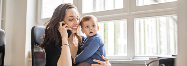 Career woman on phone at work while holding her baby who sits on her desk. 