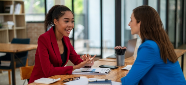 Two female employees discuss work facing each other at a table. 