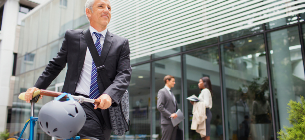 Man bikes to work with other professionals behind him in front of office building. 