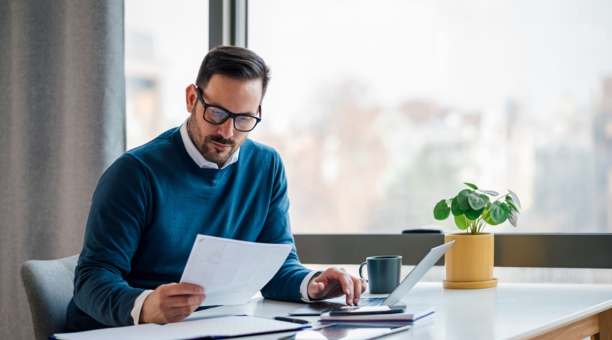 Man in blue sweater with glasses looks down at paper in his hand while sitting at a desk working. 