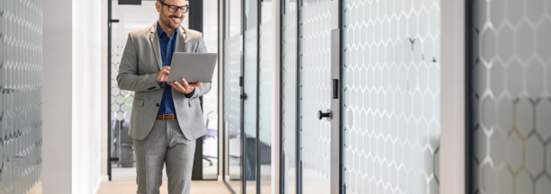 Professional man wearing glasses holding a laptop smiles and walks down hallway at work.