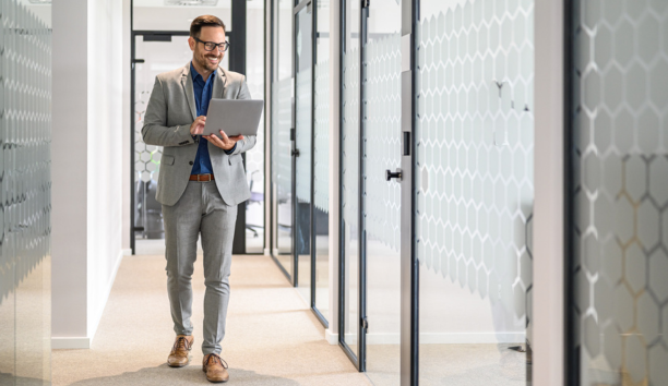 Professional man wearing glasses holding a laptop smiles and walks down hallway at work.