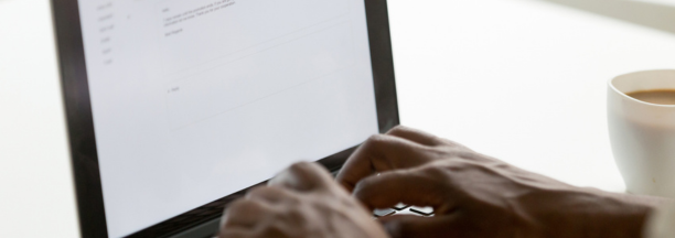 Hands of a person on a laptop keyboard writing an email with a cup of coffee next to them.