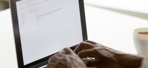 Hands of a person on a laptop keyboard writing an email with a cup of coffee next to them.