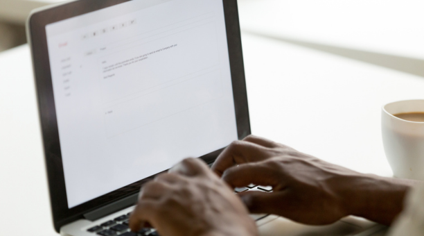 Hands of a person on a laptop keyboard writing an email with a cup of coffee next to them.