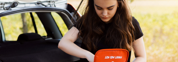 Young woman holding a first aid kit standing next to her car truck in front of a field.