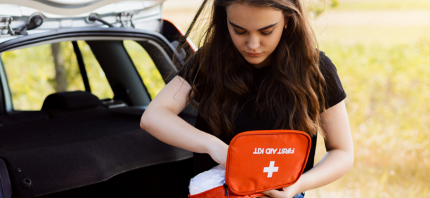 Young woman holding a first aid kit standing next to her car truck in front of a field.