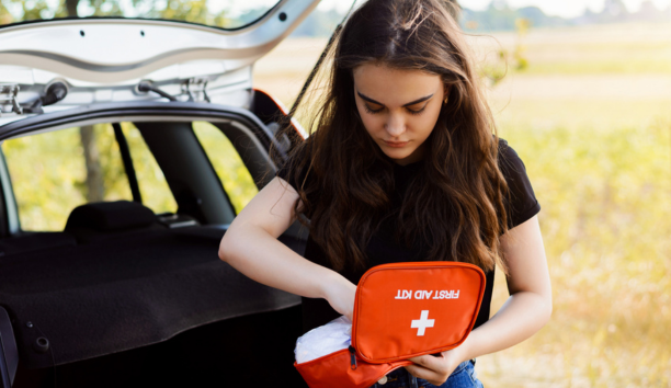 Young woman holding a first aid kit standing next to her car truck in front of a field.